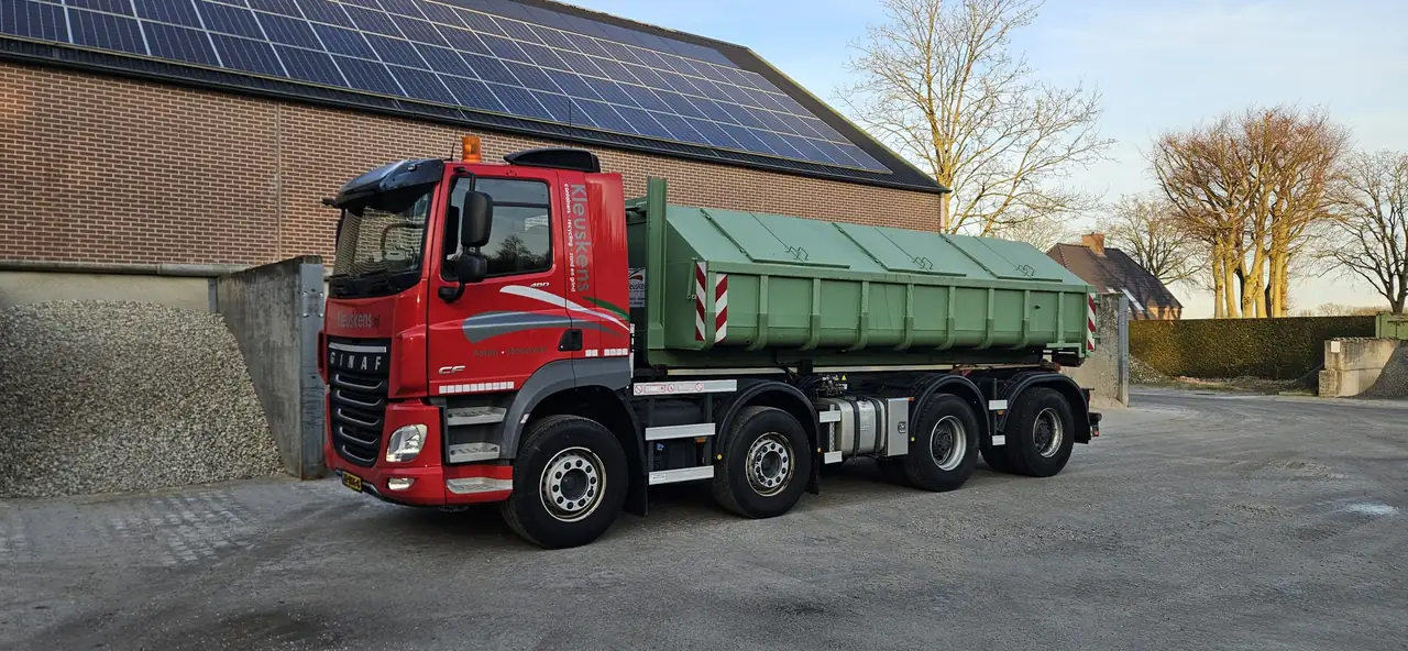 Rode GINAF-haakarmwagen van Kleuskens met een afgedekte groene haakarmcontainer naast een hoop grind