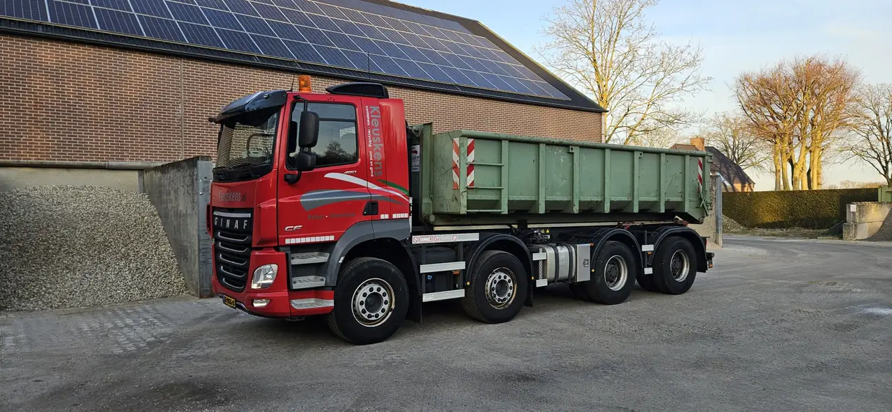 Rode haakarmwagen van Kleuskens met een open groene haakarmcontainer, voor een bakstenen loods met zonnepanelen