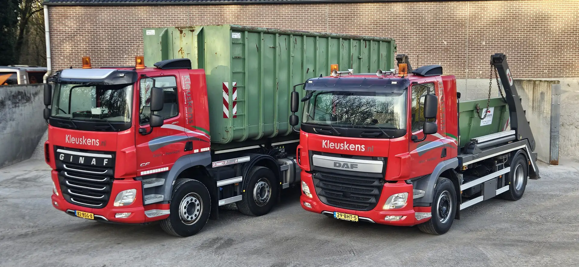 Een rode haakarmwagen en een rode portaalarmwagen van Kleuskens naast elkaar op een betonnen terrein, elk met een groene container