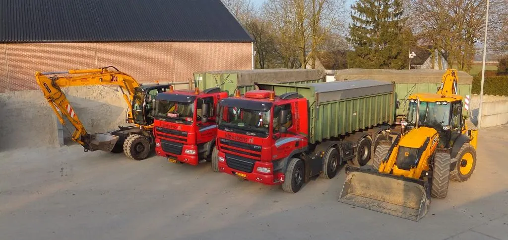 Het wagenpark van Kleuskens in de avondzon: een gele graafmachine op banden, twee rode vrachtwagens met groene containers en een gele graaflaadcombinatie
